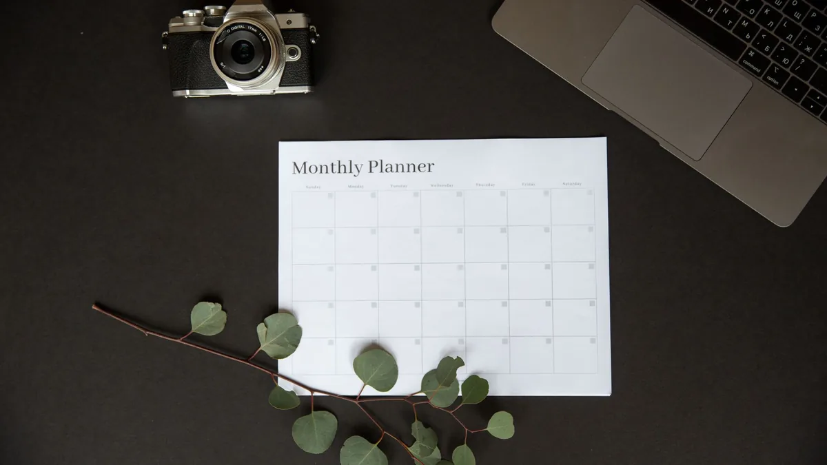 Flat lay of a monthly planner beside a camera, laptop and eucalyptus sprig
