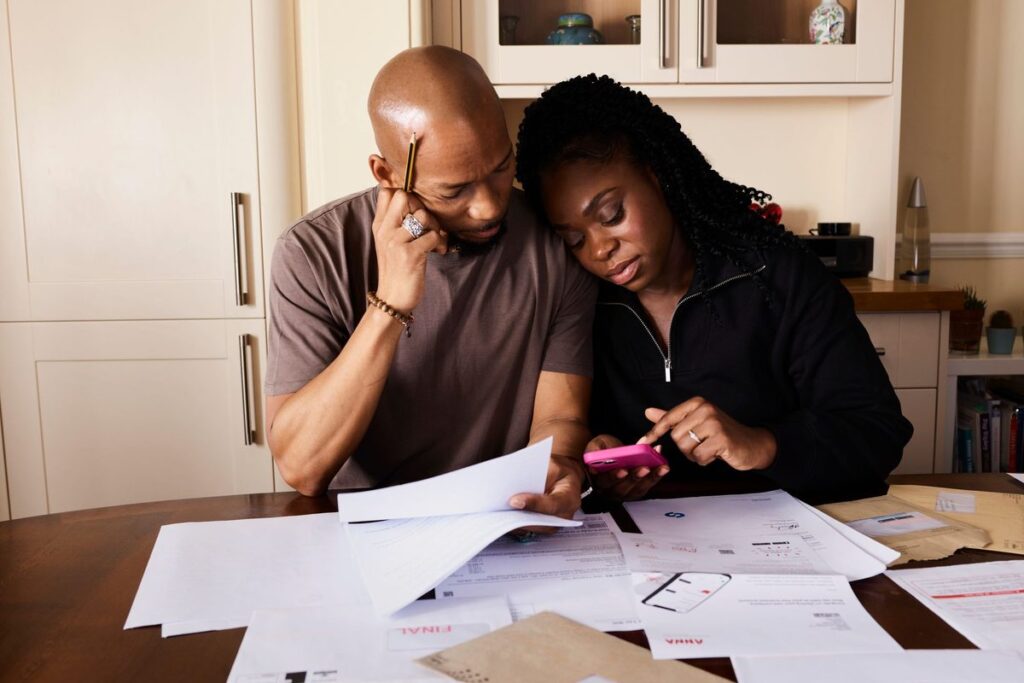 Couple sitting at a kitchen table with bills and paperwork, working out their household budget together on a phone
