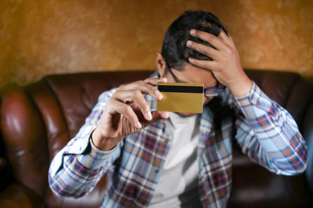 Worried man on a sofa holding a credit card with his head in his hand, thinking about his debts