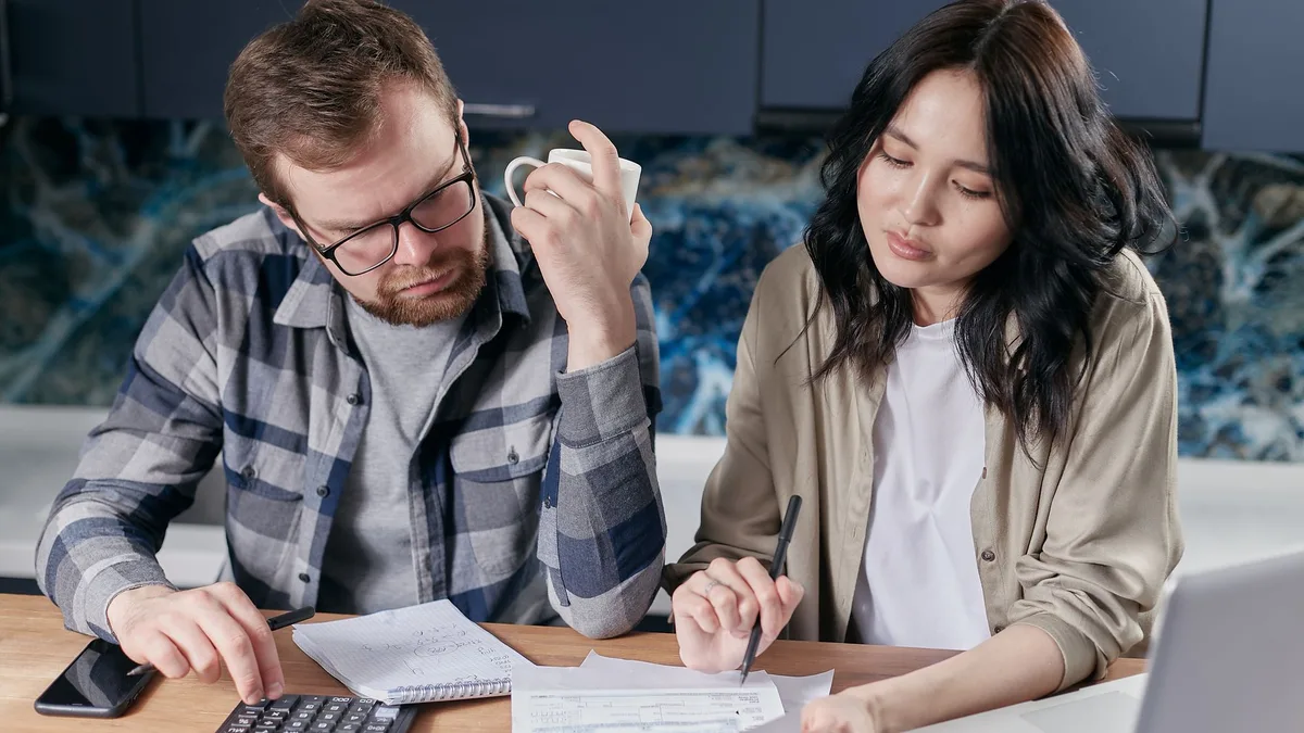Couple at a kitchen table reviewing household bills with a calculator, paperwork and a laptop