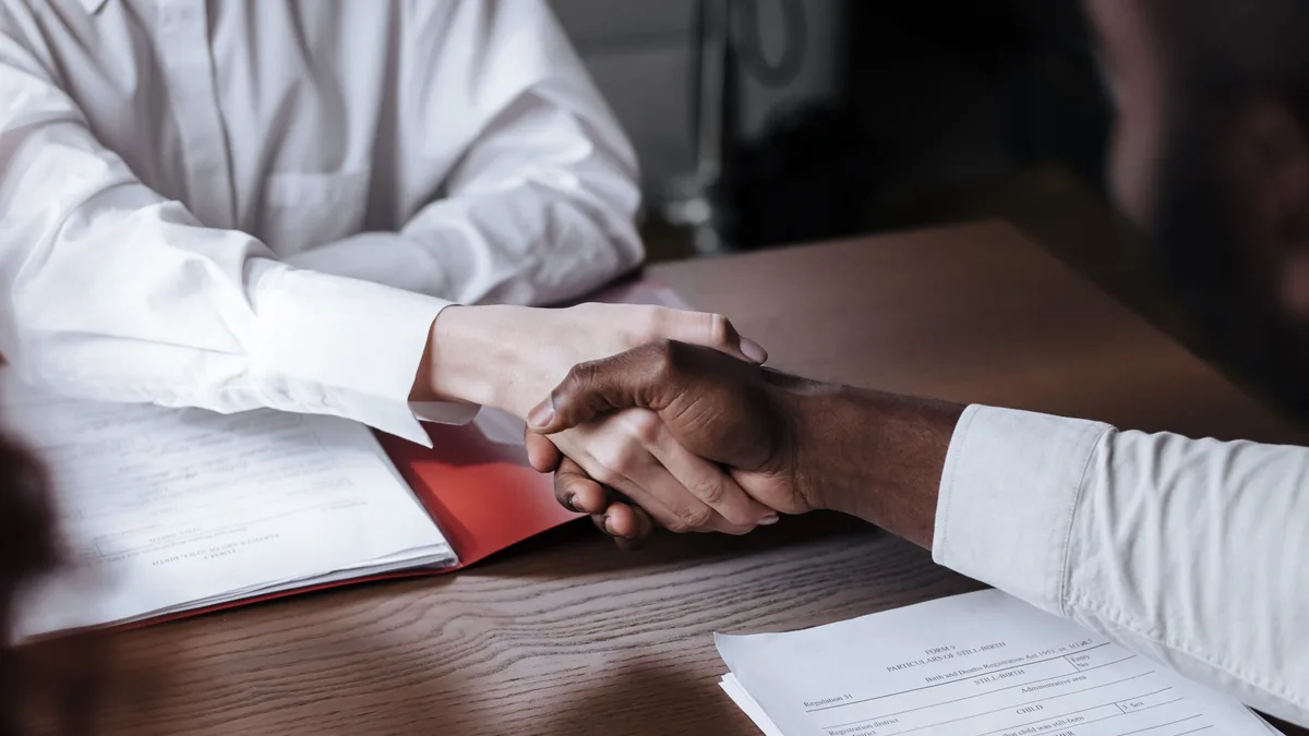 Two people shaking hands over documents across an office desk