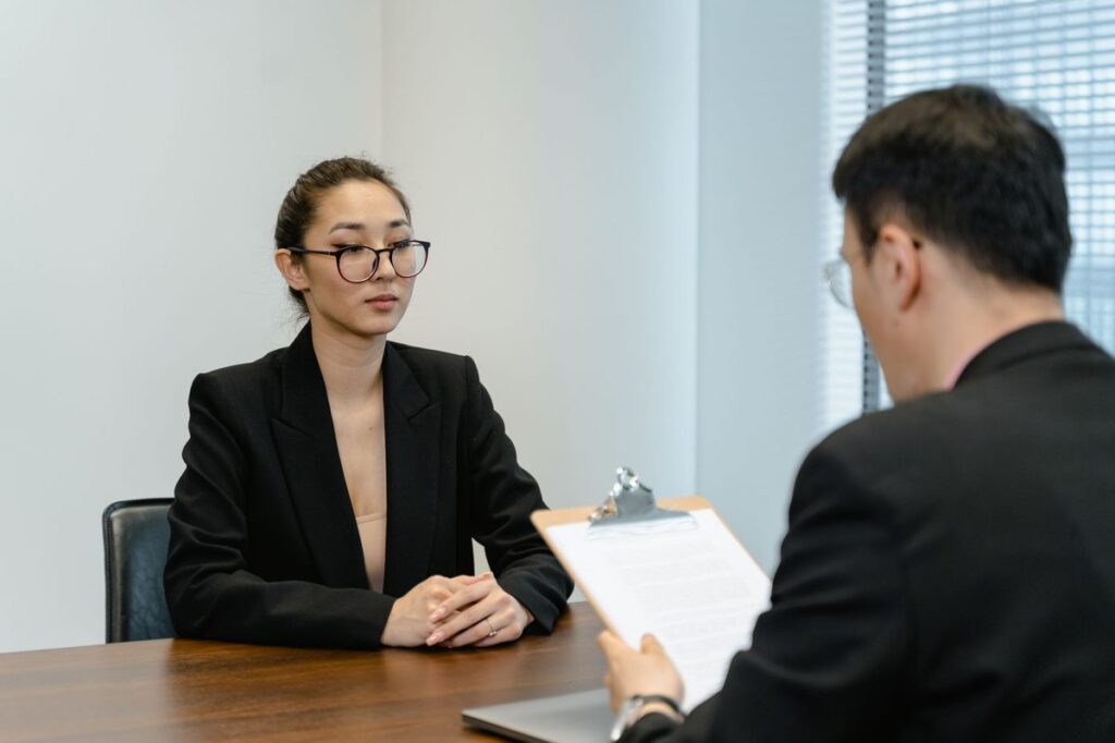 Job candidate in a black blazer sitting across a table from an interviewer holding a clipboard