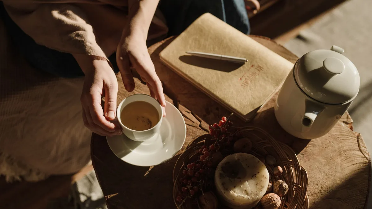 Hands holding a cup of tea beside a notebook and teapot in warm morning light