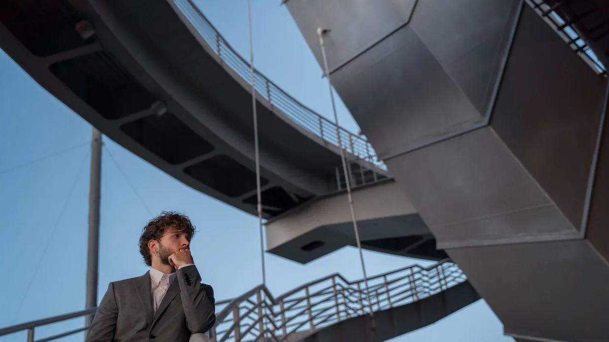 Young man in a suit standing beneath a large modern bridge, deep in thought
