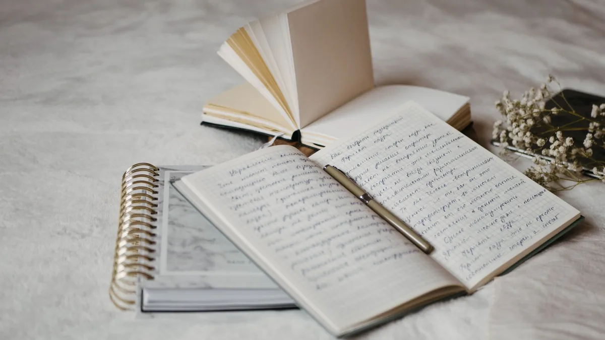 Open notebooks and a pen on a bed beside dried flowers