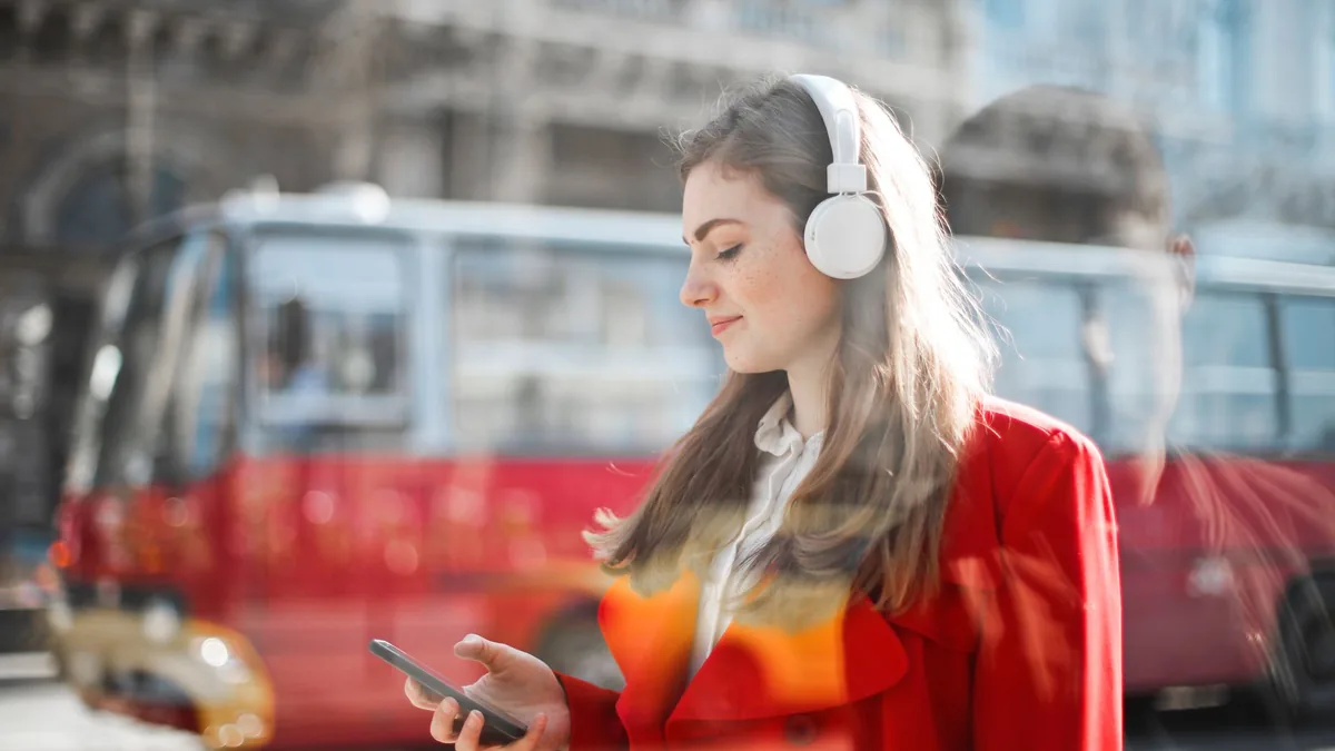 Young woman wearing headphones using her smartphone on a city street with a red bus behind her