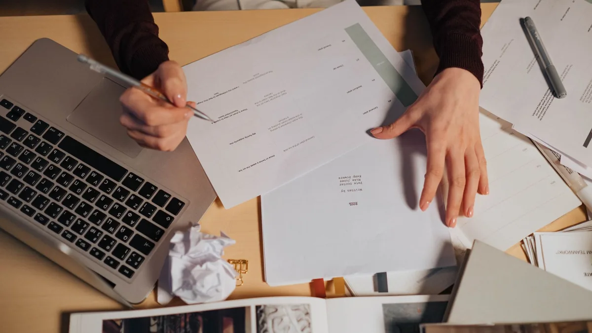Freelancer writing notes beside a laptop on a desk covered in papers