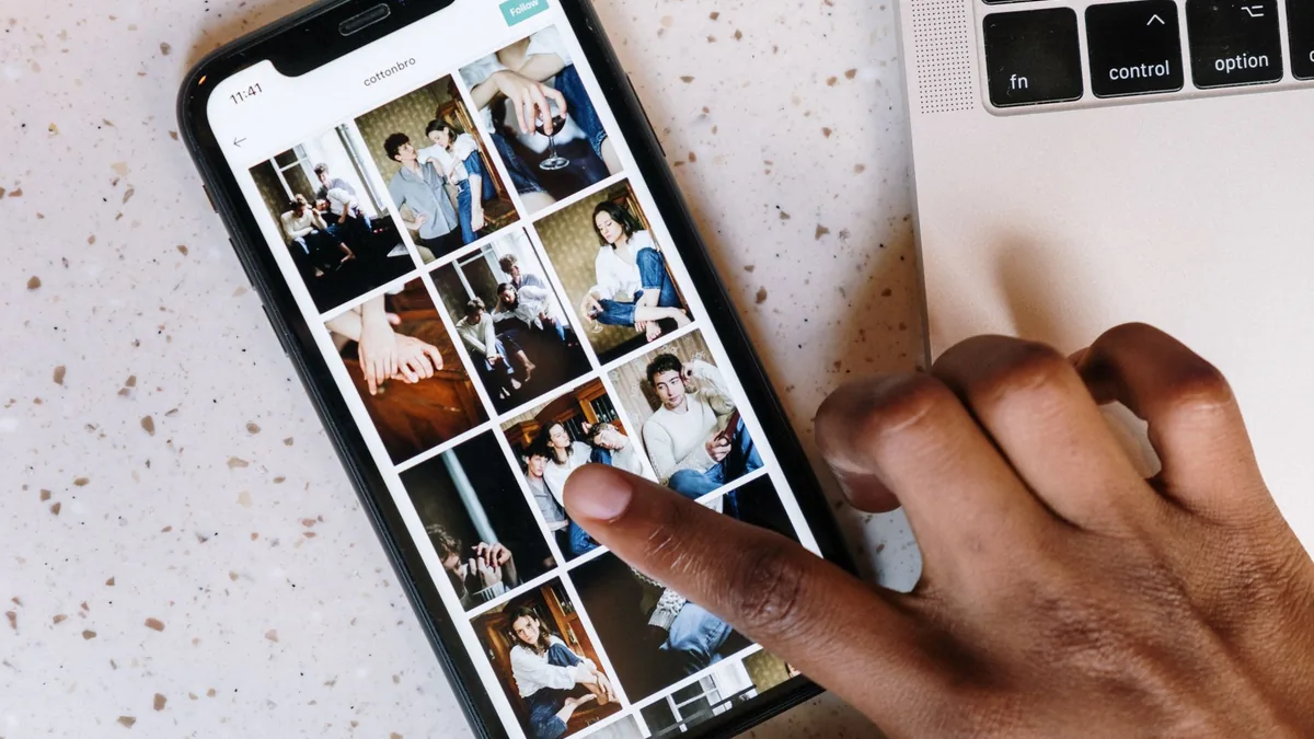 Close-up of a finger scrolling a grid of photos on a smartphone beside a laptop
