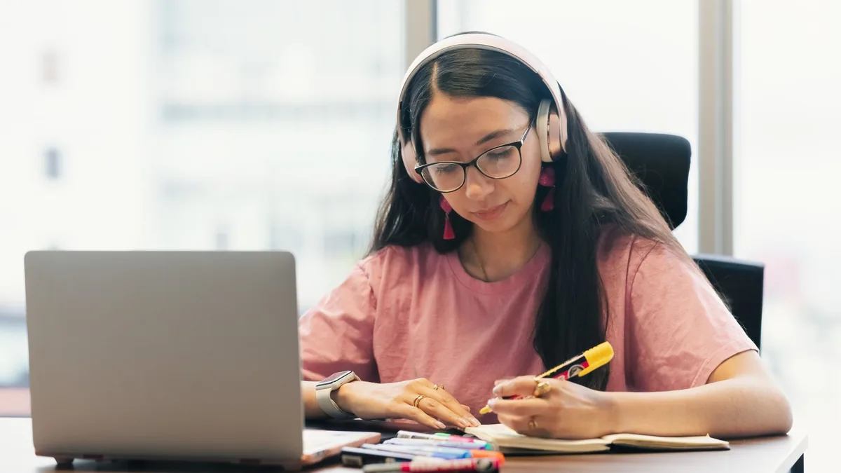 Young woman wearing headphones studying at a desk with a laptop and notebook
