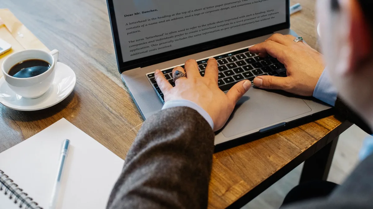 Person typing on a laptop at an office desk with a coffee, notebook and pen